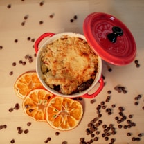A red pot contains a baked dessert with a crispy, crumbly topping. Slices of dried orange and scattered chocolate chips surround the pot on a wooden table.