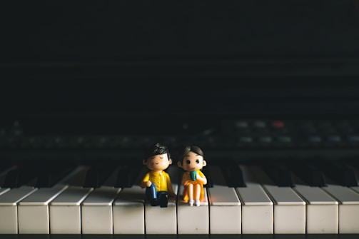Two small figurines are seated on the keys of a piano. One appears to be a boy wearing a yellow coat, and the other is a girl with a green dress. Both figurines have calm, content expressions with their eyes closed, set against a dark background that contrasts with the piano's white and black keys.