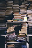 A cluttered storage area filled with stacks of books and papers, including a hand sanitizer bottle on the shelf. Books are piled on shelves and the floor, showing signs of disorder.