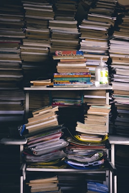 A cluttered storage area filled with stacks of books and papers, including a hand sanitizer bottle on the shelf. Books are piled on shelves and the floor, showing signs of disorder.