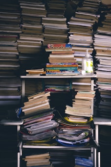 A cluttered storage area filled with stacks of books and papers, including a hand sanitizer bottle on the shelf. Books are piled on shelves and the floor, showing signs of disorder.