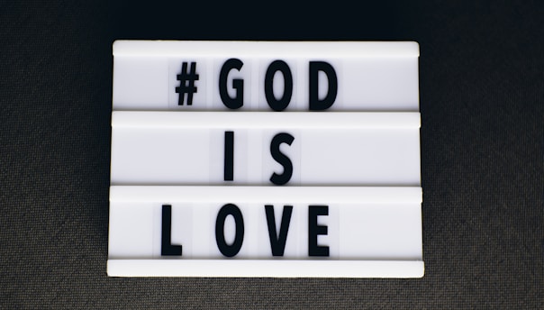 A lightbox sign featuring black uppercase letters spelling out '#GOD IS LOVE' on a white background, set against a dark surface.