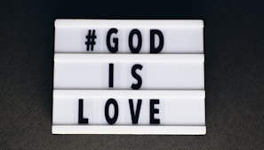 A lightbox sign featuring black uppercase letters spelling out '#GOD IS LOVE' on a white background, set against a dark surface.