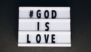 A lightbox sign featuring black uppercase letters spelling out '#GOD IS LOVE' on a white background, set against a dark surface.