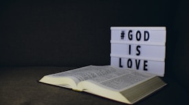 An open book lies on a dark surface with a lightbox beside it displaying the text '#GOD IS LOVE' in bold letters.