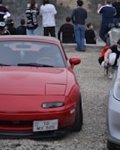 A red sports car with a license plate that reads 'AZ 10 MX 505' is parked on a gravel lot. Several people are visible in the background, standing and sitting near a railing, seemingly looking at something out of the frame. Some individuals are dressed casually, while one person is wearing a face mask and appears to be holding a helmet.