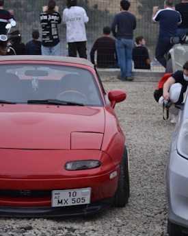 A red sports car with a license plate that reads 'AZ 10 MX 505' is parked on a gravel lot. Several people are visible in the background, standing and sitting near a railing, seemingly looking at something out of the frame. Some individuals are dressed casually, while one person is wearing a face mask and appears to be holding a helmet.