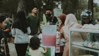 A group of people gathered around a street food stall, engaged in purchasing or observing the items available. In the center, a large container filled with a pink beverage is prominently displayed with green scoops, while various individuals are interacting with the vendor. There are trees in the background, suggesting an outdoor setting.