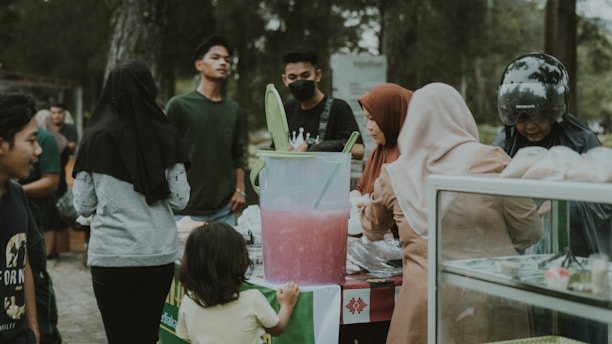 A group of people gathered around a street food stall, engaged in purchasing or observing the items available. In the center, a large container filled with a pink beverage is prominently displayed with green scoops, while various individuals are interacting with the vendor. There are trees in the background, suggesting an outdoor setting.