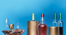 An arrangement of bottles and glasses is set against a bright blue background. The scene includes wine bottles with colored tops, glass wine glasses, a decorative bowl with a marbled pattern, and metallic vases or containers.