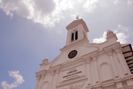 A white historic church building with classical architectural elements such as columns and intricate carvings. The facade features a large circular window, and a tall bell tower rises from the center, topped with a cross. The sky in the background is partly cloudy, with filtered sunlight creating a serene atmosphere.