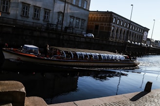 A canal tour boat with a glass roof is navigating through a calm waterway beside old, industrial-style buildings in a city setting. The sunlight reflects softly on the water, casting gentle shadows from the streetlamps along the canal.