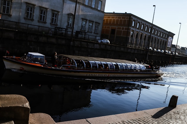 A canal tour boat with a glass roof is navigating through a calm waterway beside old, industrial-style buildings in a city setting. The sunlight reflects softly on the water, casting gentle shadows from the streetlamps along the canal.
