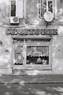A small storefront with a weathered appearance, featuring the word 'CROITORIE' prominently displayed above the door. The windows are adorned with lace curtains, and the shop sign indicates clothing repairs. Two windows and an air conditioning unit are visible above the shop, along with a satellite dish.
