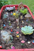 A miniature garden scene is set within a red container filled with soil. Various colorful small figurines, including gnomes and animals, are placed throughout the setting. Tiny houses and decorative plants like succulents add a whimsical touch to the scene, which appears playful and imaginative. Brightly colored artificial flowers contribute to the vibrant and fanciful atmosphere.