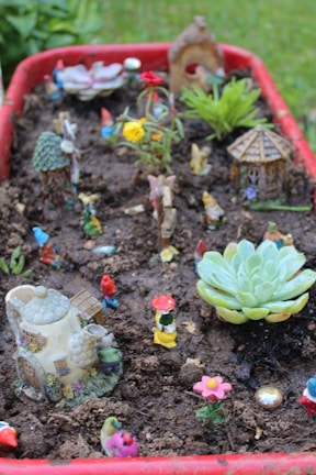 A miniature garden scene is set within a red container filled with soil. Various colorful small figurines, including gnomes and animals, are placed throughout the setting. Tiny houses and decorative plants like succulents add a whimsical touch to the scene, which appears playful and imaginative. Brightly colored artificial flowers contribute to the vibrant and fanciful atmosphere.