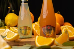 Two bottles of Sanpellegrino drinks placed on a wooden surface are surrounded by sliced lemons and oranges. The setting is vibrant, showcasing the citrus fruits prominently. The bottles have distinct labels, and the image composition suggests a fresh and natural theme with an artistic touch of blurred background.