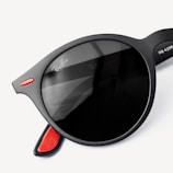 A close-up view of black, round sunglasses with a hint of red accent on the temples. The lens features subtle reflections, and there is a brand logo visible on the lens edge.