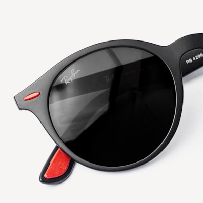 A close-up view of black, round sunglasses with a hint of red accent on the temples. The lens features subtle reflections, and there is a brand logo visible on the lens edge.