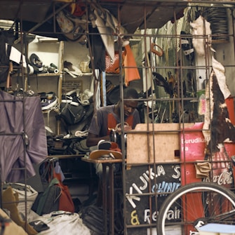 A man works in a small, cluttered repair shop filled with various tools, materials, and spare parts. Shoes and other items are scattered around, with a focus on repairs and craftsmanship. The setting is rustic and busy, indicating the shop's utility and frequent use.