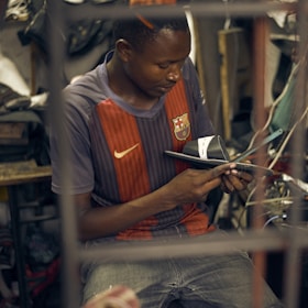 A person is concentrating intently on crafting a shoe by hand in a workshop that appears cluttered with various tools and materials. They are wearing a sports jersey and are deeply focused on their work.