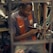 A person is concentrating intently on crafting a shoe by hand in a workshop that appears cluttered with various tools and materials. They are wearing a sports jersey and are deeply focused on their work.
