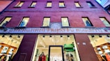 A building facade with a prominent entrance at the center. The upper part displays six windows in a symmetrical arrangement. A colorful, ornate sign in Italian decorates the area above the entrance. The interior visible through the glass doors and windows of the entrance appears to be a well-lit, stylish shop.