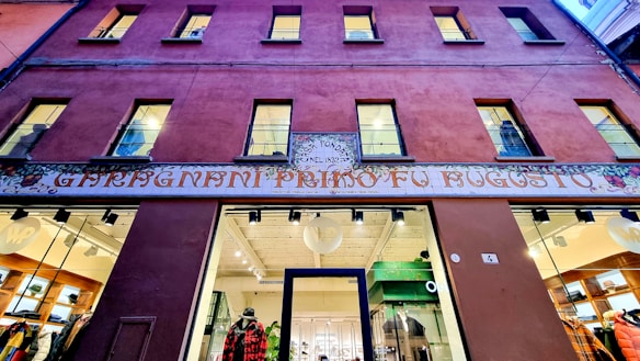 A building facade with a prominent entrance at the center. The upper part displays six windows in a symmetrical arrangement. A colorful, ornate sign in Italian decorates the area above the entrance. The interior visible through the glass doors and windows of the entrance appears to be a well-lit, stylish shop.