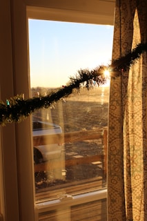 Sunlight streams through a window decorated with a festive garland, creating a warm glow. A patterned curtain hangs partially to the side, and the light outside reveals the faint outline of a car and a wooden fence.