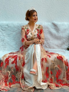A woman is sitting on a plush white sofa wearing an ornate dress embellished with red and gold patterns. She accessorizes with a small tiara and white high-heeled sandals. Her hair is styled up, and she appears poised and elegant.