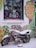 A vintage motorcycle covered in decorative beads and a layer of snow is parked in front of a brightly painted wall. The wall features colorful floral art and signage for 'Allan Gardens Floral Art Gallery'. A shop window displays high-heeled shoes, a Santa figure, and posters, alongside a scrolling LED sign with an incomplete message. The scene has a whimsical and artistic vibe, with vibrant colors despite the winter setting.