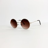 A pair of round sunglasses with a gold frame and brown-tinted lenses is placed on a light, neutral background. The thin arms of the glasses extend elegantly, suggesting a lightweight design.
