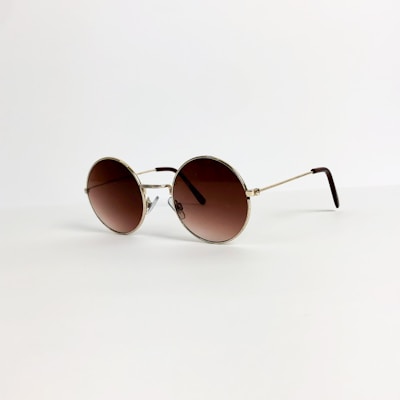 A pair of round sunglasses with a gold frame and brown-tinted lenses is placed on a light, neutral background. The thin arms of the glasses extend elegantly, suggesting a lightweight design.
