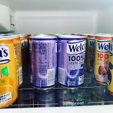 A variety of juice cans are neatly arranged on a refrigerator shelf. The visible flavors include grape, apple, and orange juice, all by the brand Welch's. The cans prominently display labels indicating 100% juice with bold colors and clear nutritional information on the side.