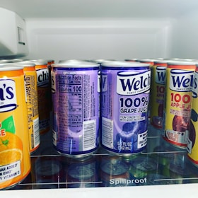 A variety of juice cans are neatly arranged on a refrigerator shelf. The visible flavors include grape, apple, and orange juice, all by the brand Welch's. The cans prominently display labels indicating 100% juice with bold colors and clear nutritional information on the side.