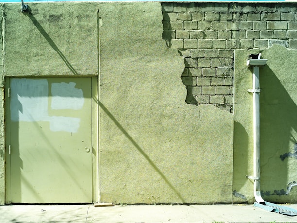 A weathered wall with peeling paint reveals underlying brickwork. The surface shows signs of wear with a few cracks and patches. A closed door is painted in a similar shade as the wall, with areas of uneven paint coverage. An external drainpipe runs vertically along the right side, casting a shadow on the textured wall.