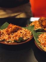 A serving of rice dish plated on green banana leaves with a piece of fried chicken on top. The rice appears spiced and colorful, indicative of biryani or a similar dish. It is presented on a dark brown plate, and the background is softly focused, suggesting a dining setting.