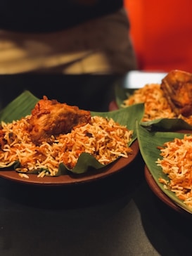 A serving of rice dish plated on green banana leaves with a piece of fried chicken on top. The rice appears spiced and colorful, indicative of biryani or a similar dish. It is presented on a dark brown plate, and the background is softly focused, suggesting a dining setting.