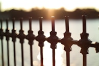 A metal fence with decorative elements is shown in the foreground. The background reveals a bright sunset over a body of water, with sunlight reflecting off the water's surface.