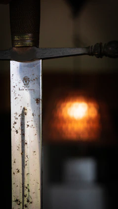 Close-up of a hand-forged sword resting beside vintage car parts on a dark wooden workbench, lit by warm brass lighting.