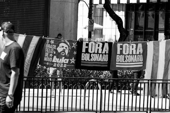 Black and white image of people standing behind a row of protest banners and flags. The banners display political messages, including 'Lula 2022' and 'Fora Bolsonaro'. A man in the foreground wears a shirt and cap, and there are trees and urban elements in the background.