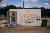 A mural featuring various cartoon characters is painted on a large white wall. Beside it, a metal cage filled with recyclable cans and a brightly painted flower stands on the ground. A green trash bin is nearby. The background is an outdoor setting with grassy areas and a partly cloudy sky.