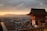A traditional Japanese temple stands in the foreground with its intricate, red wooden architecture. Behind it, a bustling city stretches out toward distant mountains under a dramatic sunset sky. Crowds of people fill the area around the temple, indicating a popular tourist spot.