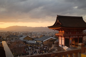 A traditional Japanese temple stands in the foreground with its intricate, red wooden architecture. Behind it, a bustling city stretches out toward distant mountains under a dramatic sunset sky. Crowds of people fill the area around the temple, indicating a popular tourist spot.