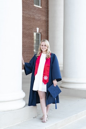 A person wearing a blue graduation gown over a white dress, standing near large white columns of a brick building. The individual is smiling, holding a blue mortarboard cap, and wearing a red stole with an emblem.