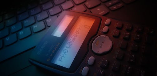 A close-up view of a scientific calculator placed on a laptop keyboard. The display screen of the calculator shows the word 'PHOTOGRAPHY' and has a soft glow contrasting with the darker surrounding keys. The lighting gives a slightly moody and dim ambiance.