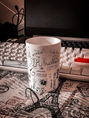A white mug with black text and graphics is placed on a surface with a patterned cloth. The mug features various words and imagery related to a superhero theme. In the background, a white computer keyboard and a partially visible open laptop are present. Cables and a headset are also visible, adding to the workspace setting.
