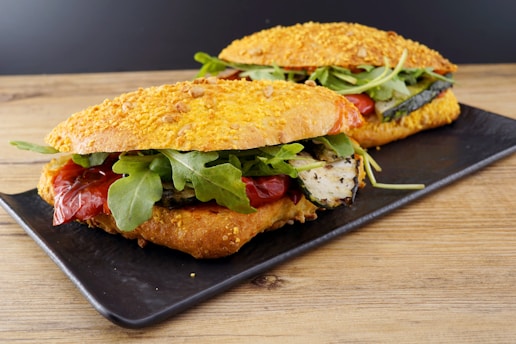 Two sandwiches on a rectangular black plate rest on a wooden surface. The bread appears to be a cheese-crusted ciabatta or similar type. Inside the sandwiches, there are grilled zucchini slices, roasted red peppers, and fresh arugula leaves, creating a vibrant and appetizing presentation.