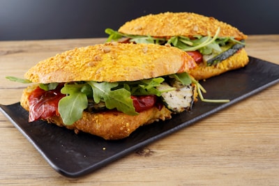 Two sandwiches on a rectangular black plate rest on a wooden surface. The bread appears to be a cheese-crusted ciabatta or similar type. Inside the sandwiches, there are grilled zucchini slices, roasted red peppers, and fresh arugula leaves, creating a vibrant and appetizing presentation.