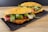 Two sandwiches on a rectangular black plate rest on a wooden surface. The bread appears to be a cheese-crusted ciabatta or similar type. Inside the sandwiches, there are grilled zucchini slices, roasted red peppers, and fresh arugula leaves, creating a vibrant and appetizing presentation.